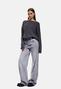 Grauer Strickpullover mit weitem Ausschnitt, kombiniert mit hellgrauen, weit geschnittenen Jeans. Ein schwarzer Gürtel und klobige schwarze Schuhe vervollständigen den Look.