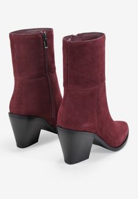 Botines de ante burdeos con punta cuadrada y cremallera en el lateral. Presentan un tacón negro robusto para mayor estabilidad.