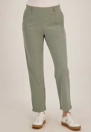 Vrouw draagt olijfgroene rechte broek met zijzakken en witte sneakers met rubberen zolen, staand voor een neutrale achtergrond.