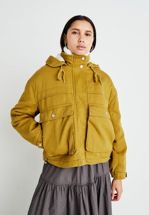 Yellow jacket with a hood, textured fabric, two front pockets, drawstring adjustments, worn over a gray ruffled dress.