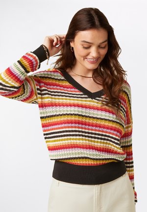 Vrouw glimlacht, draagt een kleurrijke gestreepte gebreide trui met een zwarte V-hals en boorden, en crèmekleurige broek, staand tegen een effen achtergrond.