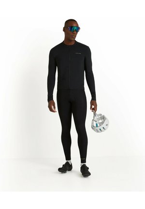 Completo da ciclismo nero con maniche lunghe e zip completa, abbinato a collant neri. Il modello tiene un casco bianco e indossa occhiali da sole riflettenti.