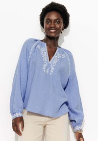 Blouse bleu clair à manches longues en tissu texturé avec un décolleté en V, ornée de motifs floraux brodés en blanc le long de l'encolure et des manches.