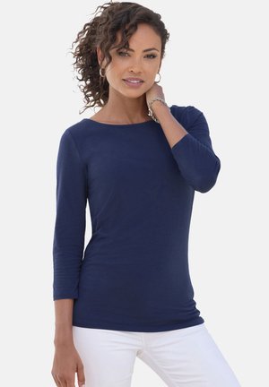 T-shirt a maniche lunghe blu navy con scollatura a barca e una texture morbida, abbinato a pantaloni bianchi. Presenta un design aderente e una leggera drappeggiatura.