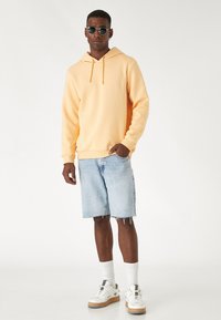 Jasnopomarańczowa bluza z kapturem ze sznurkiem, w zestawie z jasnoniebieskimi szortami dżinsowymi. Model nosi białe sneakersy i czarne okulary przeciwsłoneczne. Prosty, casualowy design.