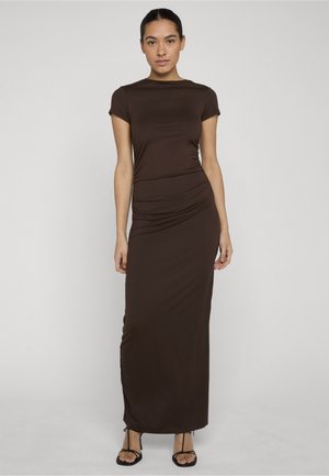 Vrouw met donker haar in een lange, nauwsluitende bruine jurk met korte mouwen en zwarte sandaaltjes met bandjes, staand tegen een effen lichte achtergrond.