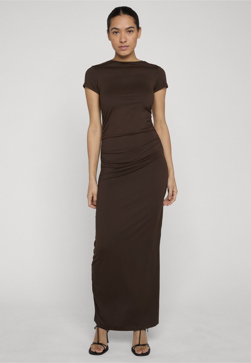 Femme aux cheveux foncés portant une robe marron longue et cintrée à manches courtes et des sandales noires à brides, debout devant un fond clair uni.