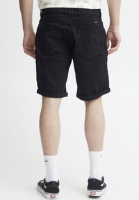 Shorts en denim noir avec des revers, comportant des poches arrière, portés avec des baskets noires et des chaussettes blanches à la cheville. Design et texture minimalistes.