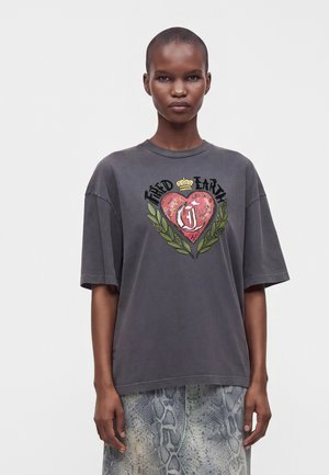 Mujer con el pelo corto que lleva una camiseta holgada de color gris oscuro con un gráfico de un corazón y "Fated Earth" y pantalones con estampado de serpiente.