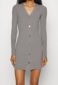 Robe en maille grise côtelée avec un col en V, des manches longues et une fermeture à boutons sur le devant. Elle présente une fente haute à la cuisse et des détails de boutons métalliques.