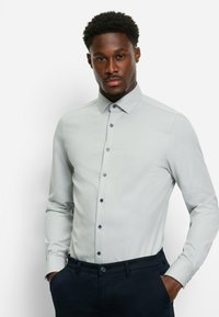 Hellgraues Hemd mit Knopfleiste, dezentem Muster, langen Ärmeln und marineblauen Akzenten an den Knöpfen. Kombiniert mit dunklen Hosen.