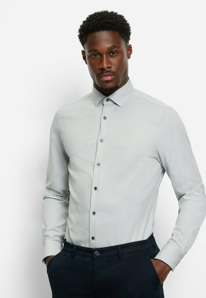 Light grey button-up shirt with a subtle pattern, long sleeves, and navy accents on buttons. Paired with dark trousers.