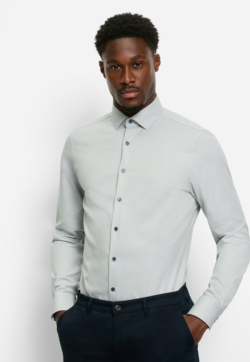 Hellgraues Hemd mit Knopfleiste, dezentem Muster, langen Ärmeln und marineblauen Akzenten an den Knöpfen. Kombiniert mit dunklen Hosen.