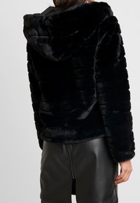 Giacca nera in pelliccia sintetica con cappuccio, caratterizzata da una texture a coste e una vestibilità rilassata. Indossata sopra una gonna in pelle aderente con dettaglio della zip.
