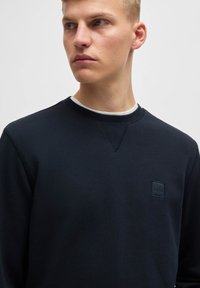 Granatowy sweter z okrągłym dekoltem wykonany z miękkiego materiału, z małą, teksturowaną naszywką z logo na piersi i widocznymi przeszyciami.