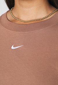 Brązowy bawełniany t-shirt z dekoltem w serek, z białym logo Nike na piersi i gładką, teksturowaną powierzchnią.