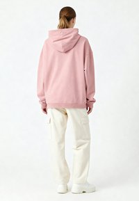 Haut à capuche oversize rose avec une capuche, des poignets et un ourlet côtelés, associé à un pantalon cargo blanc cassé avec des poches latérales et des baskets blanches.