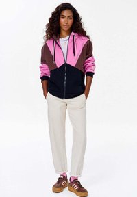 Kurtka z zapięciem na zamek z kolorowymi blokami w odcieniach różu, brązu i czerni. Połączona z kremowymi spodniami i brązowymi sneakersami z różowymi akcentami.
