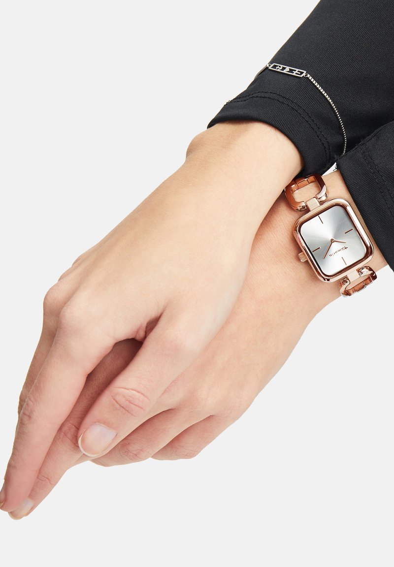 Rechteckige Uhr aus Roségold mit silbernem Ziffernblatt und vier minimalistischen Stundenmarkierungen, montiert an einem Gliederarmband, getragen an einem Handgelenk mit heller Hautfarbe.