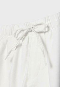 Gros plan d’un pantalon blanc à taille élastique avec un cordon noué en nœud et des coutures visibles sur la poche latérale.