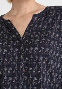 Marinblus i lätt tyg med ett vertikalt pilmönster i vitt, rött och blått. V-ringning med subtila virkdetaljer.