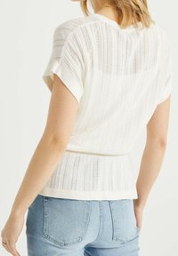 Il maglione bianco lavorato a maglia presenta una texture a coste con strisce orizzontali, maniche corte e una vita aderente. Ideale per un look casual.