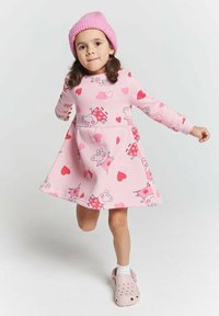 Junges Mädchen mit pinker Mütze, pinkem Kleid mit Cartoon-Schwein und Herzmustern, weißen Socken und pinken Slippern, das mit einem Bein angehoben posiert.