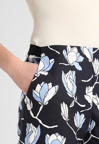Kalhoty s květinovým vzorem, které mají navy modrou základnu a modro-bílé designy magnolie. Hladká tkanina se bočními kapsami a černým pasem.