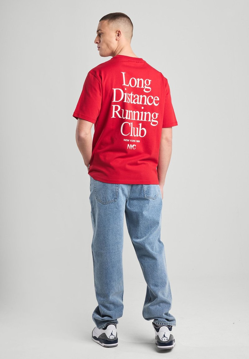 Punainen puuvillainen t-paita, jossa on valkoisella painettu "Pitkien matkojen juoksuklubi" selkäpuolella, yhdistettynä vaaleansinisiin farkkuihin ja tennareihin.