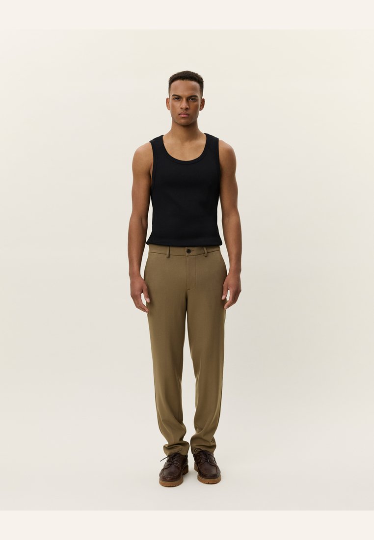 Black sleeveless tank top paired with tan pleated trousers. Brown lace-up shoes complete the outfit. Neutral backdrop.