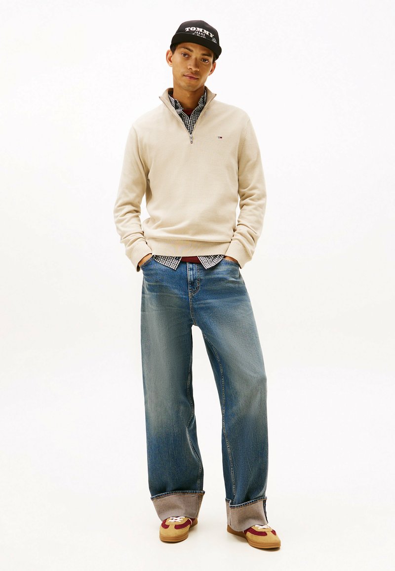 Jersey beige con cremallera, camisa de cuadros debajo, pantalones vaqueros anchos azules y calzado con detalles marrones, sobre un fondo sencillo.