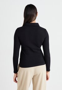 Camisa polo negra de manga larga con cuello, con un diseño ajustado y textura de canalé, llevada con pantalones beige, vista desde la espalda.