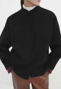 Veste en mélange de laine noire avec un col haut, fermeture à boutons et design surdimensionné. Présente deux poches avant et des accents de poignet côtelés.