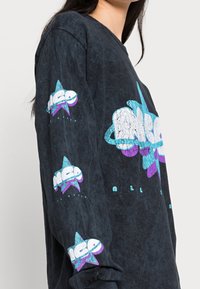 Černá košile s dlouhým rukávem, která obsahuje barevné grafické potisky hvězd a textu na rukávech a přední části, s texturovaným, vybledlým vzhledem.