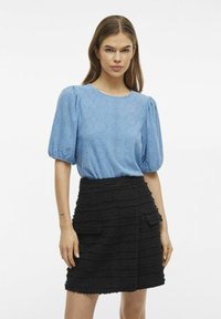 Ljusblå blus med ögonstygnsdesign och puffärmar, tillsammans med en texturerad svart kjol med lager av tyg och framfickor.