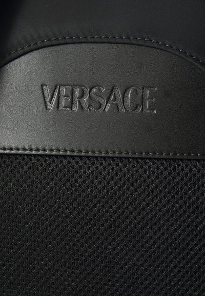 Melna teksturēta auduma ar sietu dizainu, ar izsistītu "VERSACE" logotipu melnā krāsā uz gludas ādas līdzīgas virsmas.