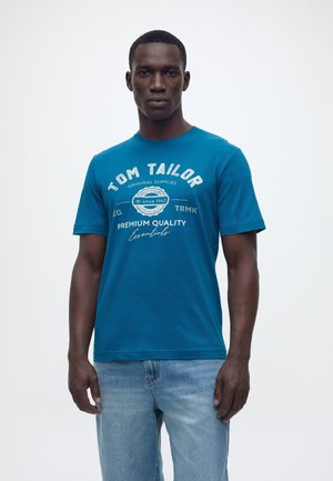 Homme portant un t-shirt teal Tom Tailor avec un texte blanc et un jean bleu clair, debout devant un fond blanc uni.
