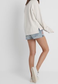 Sweat-shirt oversized beige clair avec fente latérale, associé à un short en jean bleu clair déchiré. Le modèle porte des baskets blanches à semelles texturées.
