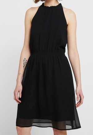 Frau trägt ein ärmelloses schwarzes Neckholder-Kleid mit elastischer Taille und transparentem Saum, das florale und geometrische Tattoos auf den Armen zeigt.