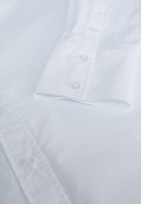Tessuto di camicia in cotone bianco con una texture liscia, dotato di un polsino con bottoni, cinque bottoni e un design pulito e classico.