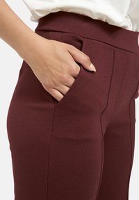 Hand in der vorderen Tasche einer hochtaillierten, burgunderfarbenen Hose, die mit einem weißen Oberteil getragen wird, wobei die Stofftextur und der Sitz deutlich sichtbar sind.