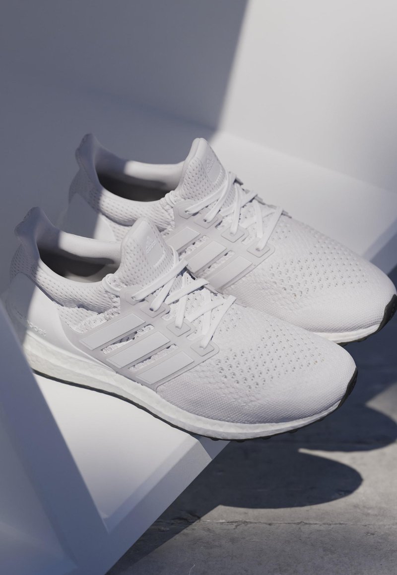 White athletic shoes feature a breathable mesh upper, textured accents, three side stripes, and a cushioned sole with a black outsole.