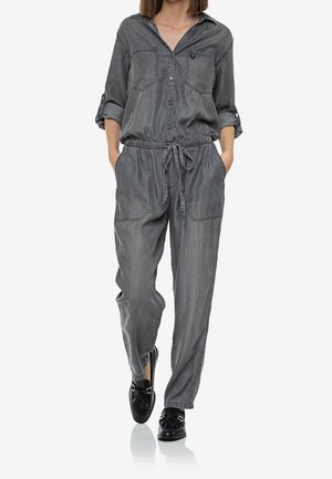Kvinde iført en løs grå denim-dragt med opsmøgede ærmer og sorte loafers, der går med hænderne i lommerne mod en hvid baggrund.
