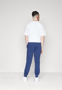 Λευκό μπλουζάκι με λαιμόκοψη, μπλε φόρμες με μανίκια στο τελείωμα, συνδυασμένα με λευκά αθλητικά παπούτσια, προβαλλόμενο από πίσω σε ουδέτερο φόντο.