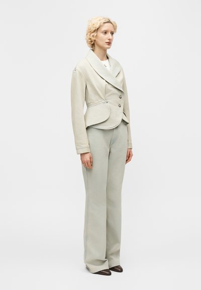 Žena s krátkymi kučeravými blond vlasmi, oblečená v svetlobéžovom priliehavom dvojradovom saku a zodpovedajúcich širokých nohaviciach, stojaca zboku.