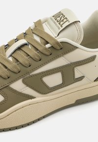Green and cream low-top sneaker with a textured rubber sole, featuring stitched accents, round laces, and a brand logo tag on the tongue.