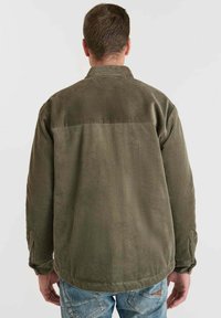 Man met zijn rug naar ons toe, gekleed in een olijfgroene corduroy jas en vervaagde blauwe jeans, tegen een effen lichte achtergrond.