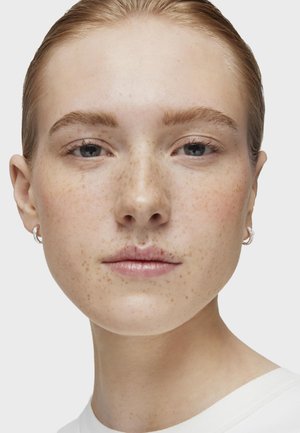 Primer plano de una cara con piel clara, pecas y maquillaje natural. Lleva pendientes de aro plateados y una camisa blanca con un escote redondo.