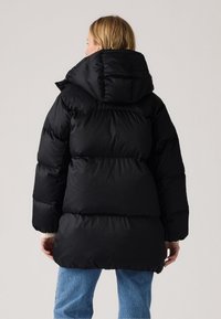 Black puffer jacket with a large hood, featuring quilted sections, a smooth texture, and a relaxed silhouette. Worn over light blue jeans.