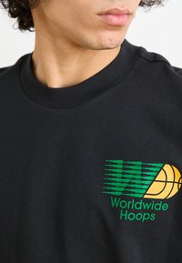Juodos medvilnės marškinėliai su žaliai geltonu grafinio dizaino atvaizdu, kuriame yra tekstas "Worldwide Hoops" ir krepšinio ikona, lygi tekstūra, apvalus iškirpimas.